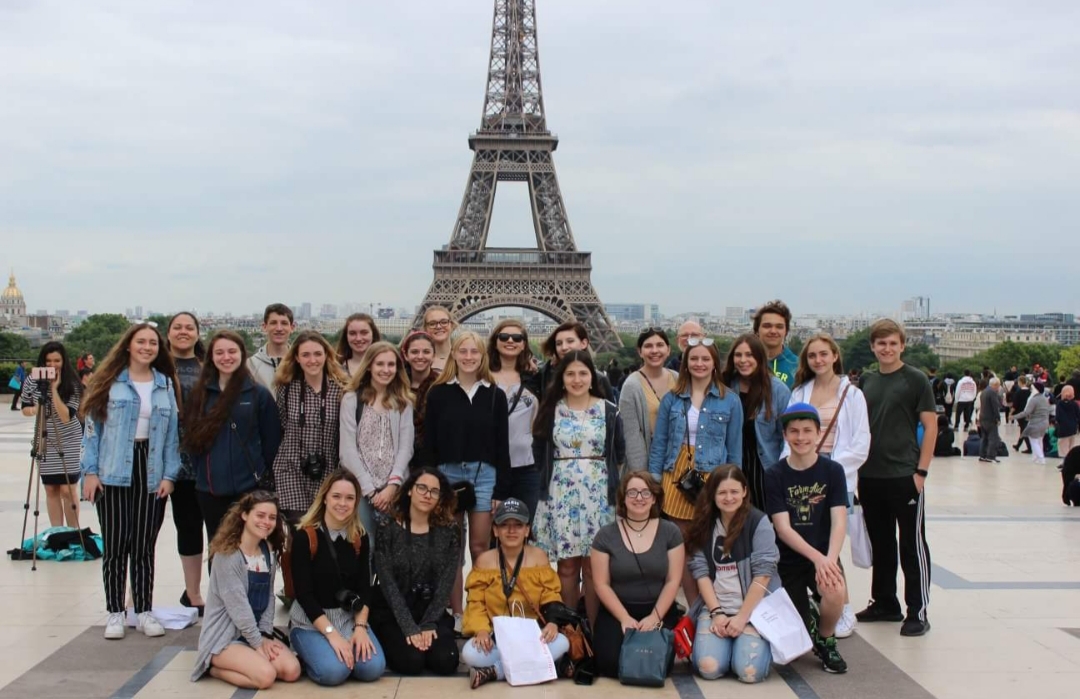 The students all stand in front of the Eiffel Tower. The tower is one of the most the most famous architectural icons of the world and is easily recognized by many people.