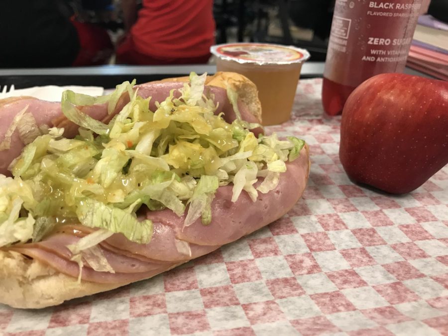 Sweet and Salty School lunches lie out on a plastic tray awaiting to be eaten. A ham and cheese hoagie, apple and two beverages were placed upon the tray in an aesthetically pleasing way to suit the students.