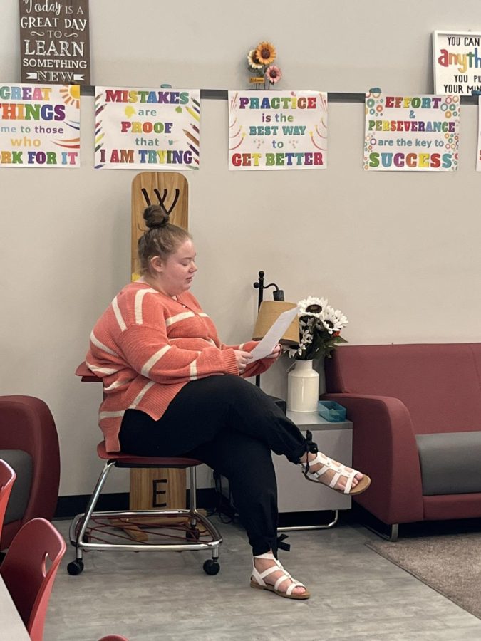 Harris is a special education teacher. She is reading the paper to her students while sitting in her mini setup with the sign and the flowers on the table next to a couch. 