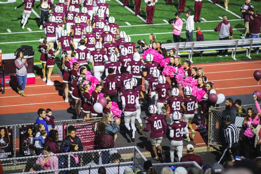 The cheerleaders raise their pom-poms up to lead in the football players to play at the breast cancer awareness game. The players are guided onto the field by pink pom-poms to support breast cancer awareness. 