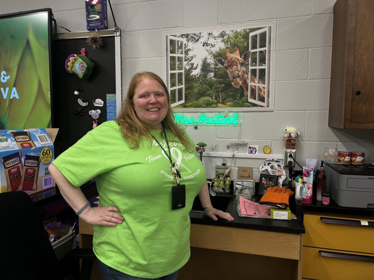 Biology+teacher+Heather+Partsch+poses+in+front+of+her+neon+sign.+The+neon+sign+is+lime+green%2C+unintentionally+the+same+color+as+the+T-shirts.+